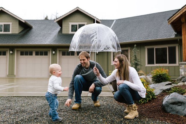 Image of mom and dad crouching down holding an umbrella over their young son. They are standing outside of their home.